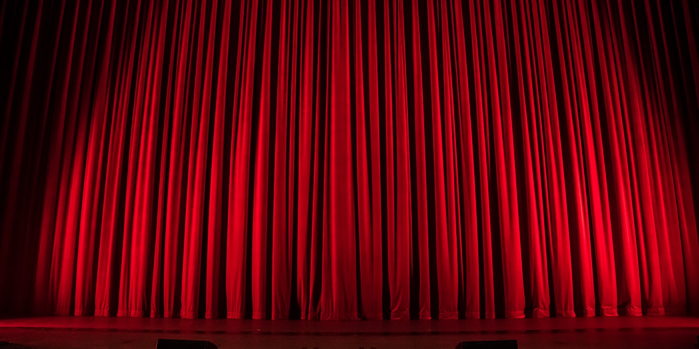 Rood theaterdoek in warm toneellicht, vlak voor de opening